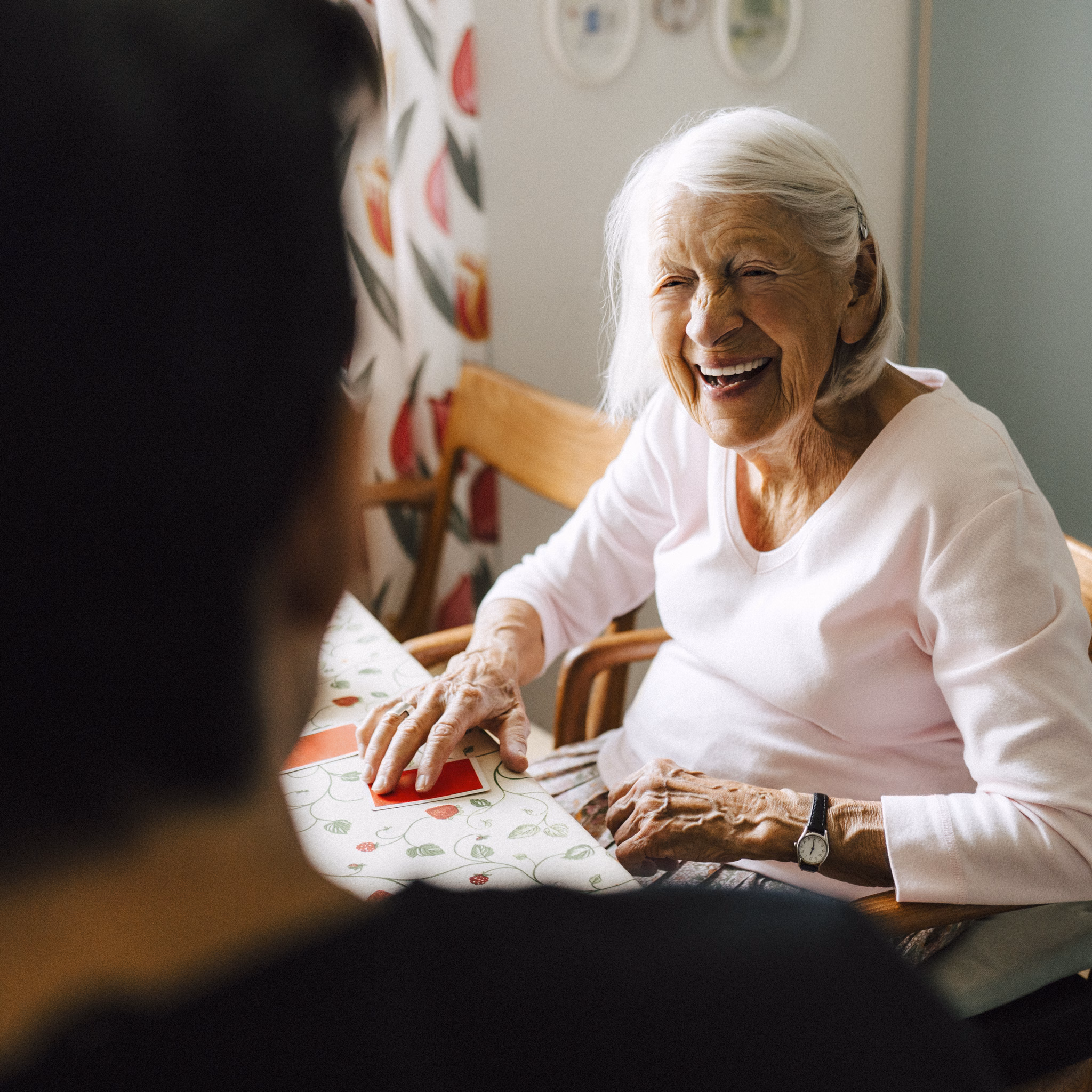 Fröhliche Seniorin sitzt am Tisch und spielt mit jüngerer Person Karten
