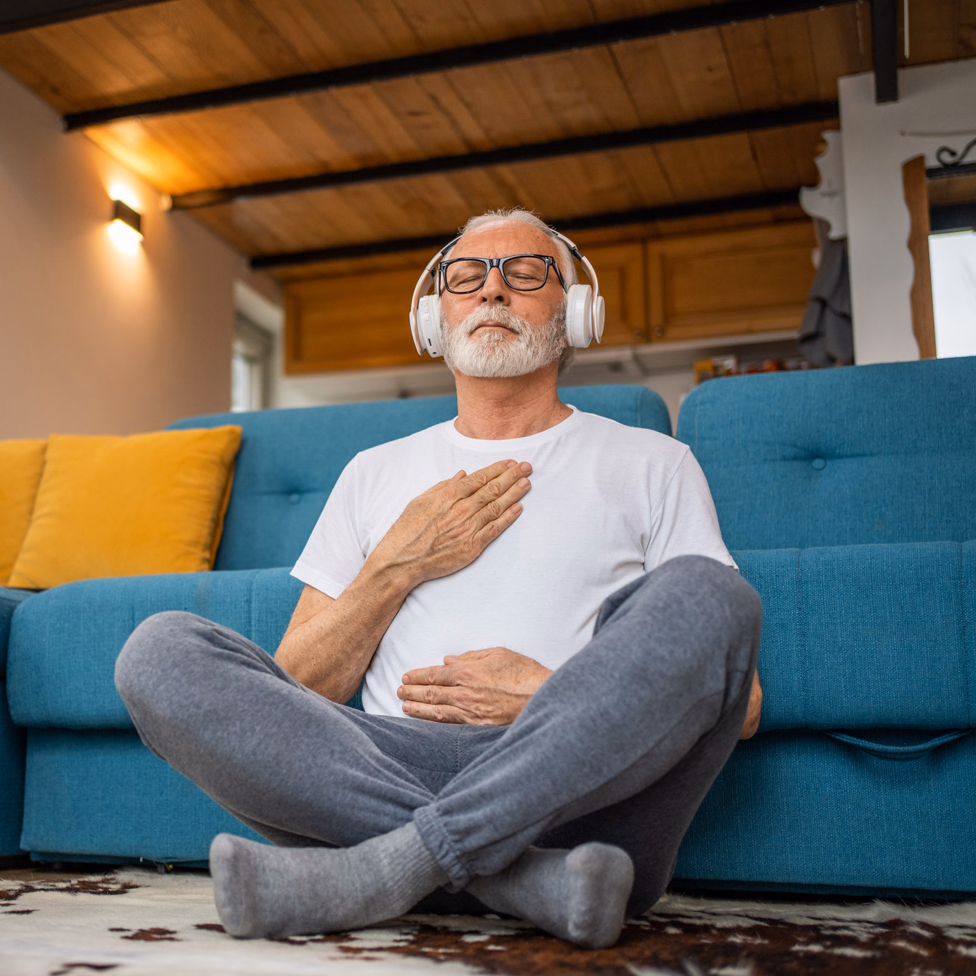 70 jähriger Mann sitzt vor Sofa und macht Atemübungen.